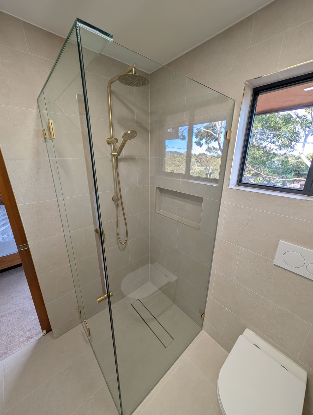 Frameless corner shower screen with a fixed return panel and hinged door, brushed brass hinges, in a beige-tiled bathroom