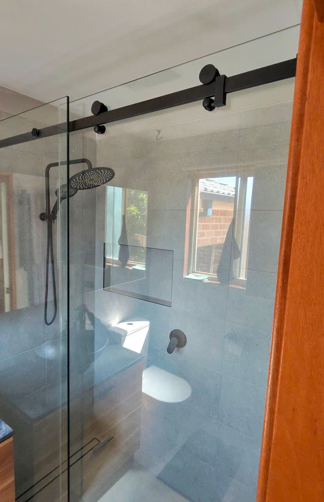 Frameless sliding shower door on a matt black top track, grey tiles and timber vanity